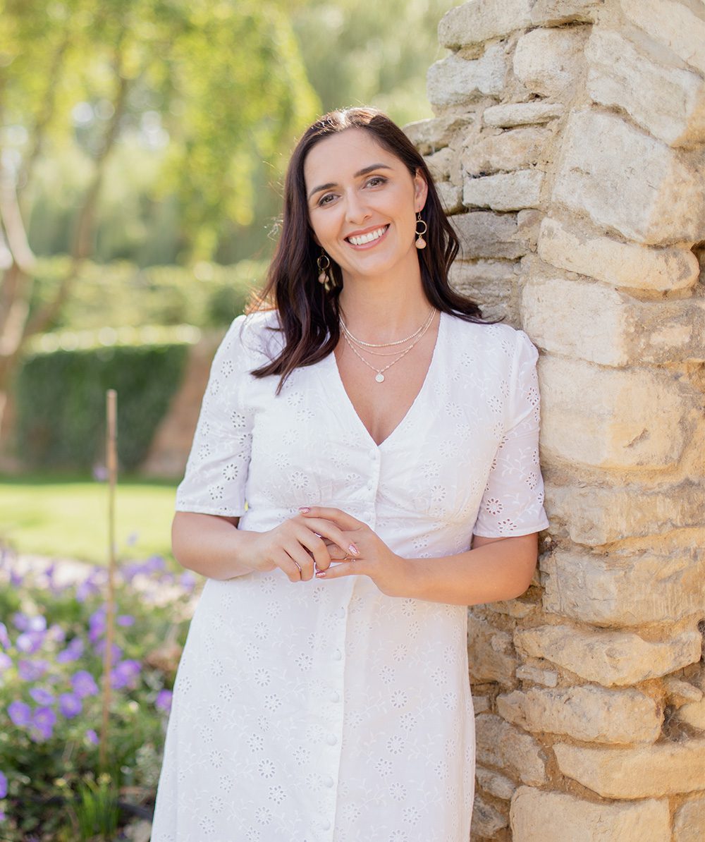 Natasha Leigh Bray leaning against a stone coloured brick archway wearing a white dress